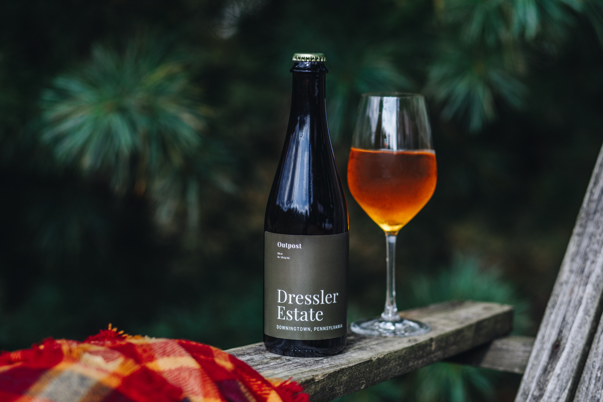 A 500 mL bottle of Dressler Estate's Outpost cider with a filled glass and a flannel blanket on the arm of an Adirondack chair.
