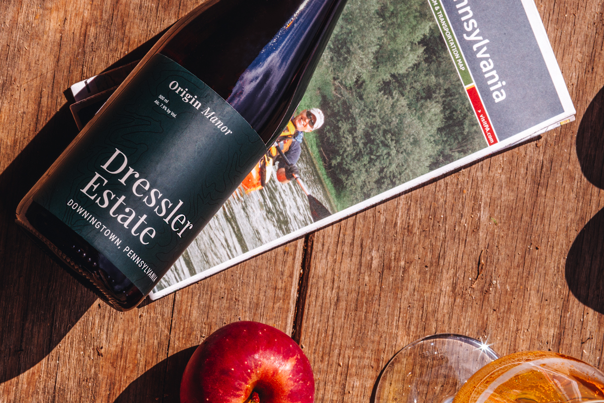 A 500 mL bottle of Dressler Estate's Origin Manor cider with a filled glass, apple and folded map of Pennsylvania on a barn floor.