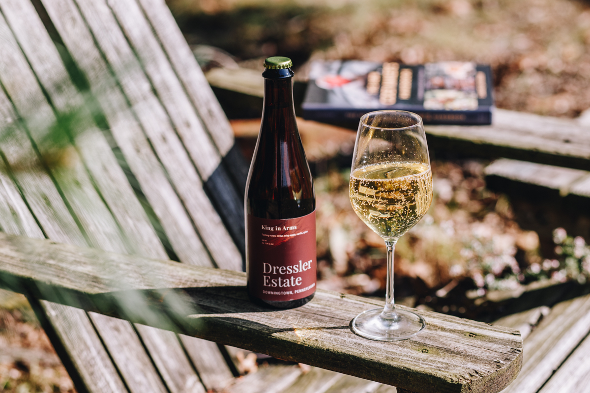 A 500 mL bottle of Dressler Estate's King in Arms cider with a filled glass on the arm of an Adirondack chair.