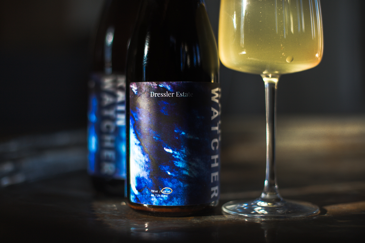 A filled glass of cider with 500 and 750 mL bottles of Dressler Estate's Rain Watcher.